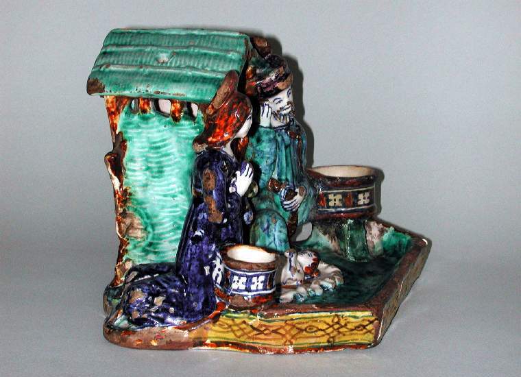 An image of Maiolica. Nativity Inkstand. Giovanni di Nicola Manzoni dal Colle (active early 16th century, Italian, Faenza, circa 1507-1516). With a Nativity group in the round at the back, and an inkwell and candlesocket on either side. The Virgin Mary and St Joseph kneel on either side of a wattle stall occupied by an ox and an ass. The infant Christ lies on a wavy-edged cloth between them. In front of the figures is a space for pens, enclosed by a low wall, decorated on the exterior with strapwork. Dark buff earthenware, tin-glazed on the upper surfaces; base streaked with glaze. Painted in dark blue, green, yellow, brownish-orange, and manganese-purple high-temperature (metallic oxide) colours, height, whole, 15.8 cm, length, whole, 21.5 cm, depth, whole, 17.1 cm, circa 1509-1510. Renaissance.