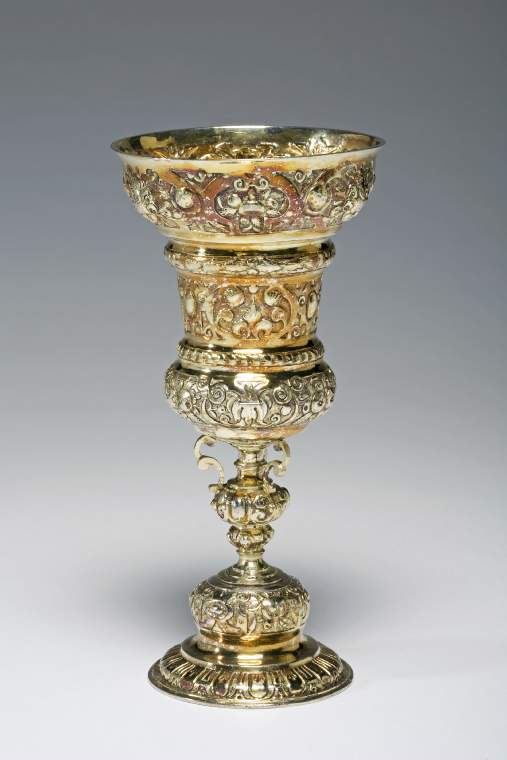 An image of Standing Cupsilver-gilt; highly domed foot and waisted bowl embossed and chased with strapwork, cherubs' heads, swags, fruit and foliage.Silver-gilt; the circular foot is stepped and highly domed in the centre.  The stem has an urn-shaped knop with three applied scrolls supporting the bowl, which is waisted with a flared rim.  The bowl, foot and knop are embossed and chased with borders of strapwork, cherubs' heads, swags, flowers and foliage on a matted ground.