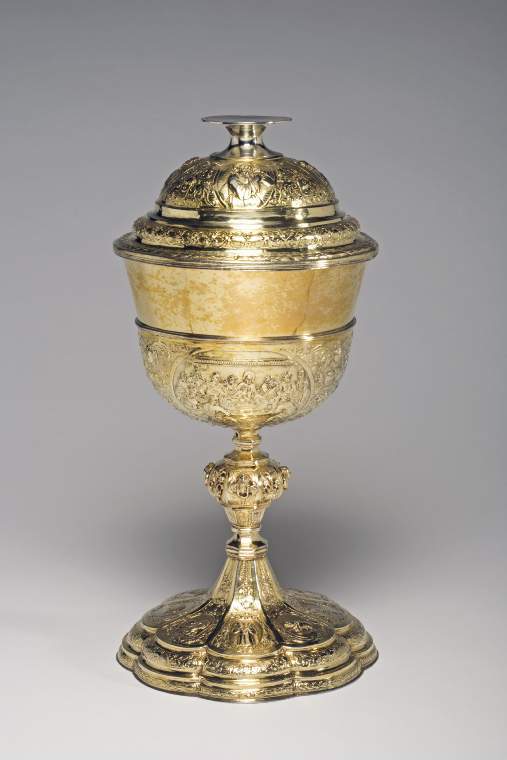 An image of Ciborium and coversilver-gilt; large bowl with three carouches of Biblical scenes, the octofoil foot and domed cover similarly decorated.Silver-gilt; the large bowl has a slightly everted rim and sits within a shallow cup, which is embossed with three oval cartouches containing vignettes of Christ at Emmaus, the Last Supper and Manna arriving from Heaven.  Between the three cartouches are formal arrangements of cherubs' heads, fruit and scrolling foliage on a striated ground.  The octagonal stem has a large central knop applied with a border of alternating cherubs' heads and lilies in urns.  The stepped spreading octafoil foot is embossed with eight oval cartouches containing Christian symbols, including the Agnus Dei,  the pelican in her piety,  the Good Shepherd, the lion of Judah and a lily for purity, a beehive, a wheatsheaf and a coat-of-arms with the initial 'W' above and 'E' and 'F' on either side.  These are surrounded by formal borders containing cherubs' heads, fruit and scrolls on a striated ground.  The stepped and domed cover is similarly decorated and is embossed with four oval cartouches depicting the Passions of Christ.  The spool-shaped knop cum foot is undecorated and has a slightly concave upper surface.