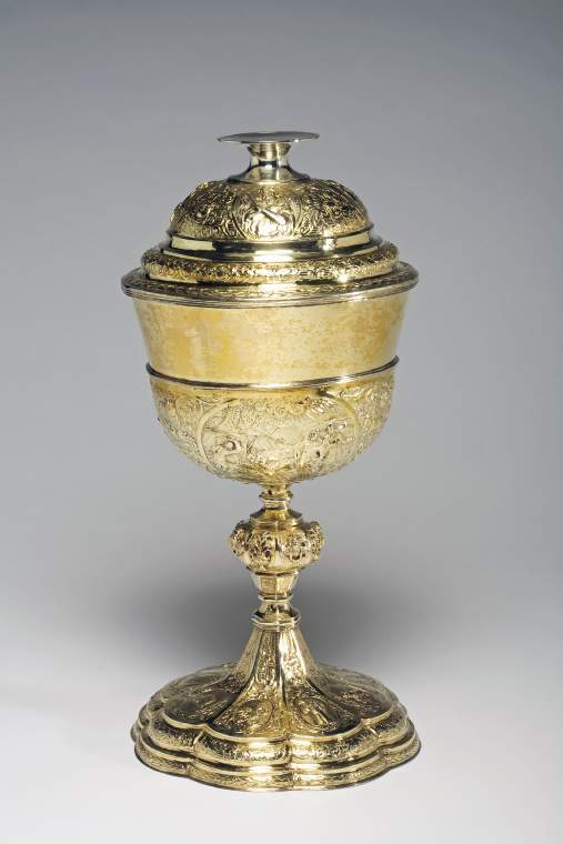 An image of Ciborium and coversilver-gilt; large bowl with three carouches of Biblical scenes, the octofoil foot and domed cover similarly decorated.Silver-gilt; the large bowl has a slightly everted rim and sits within a shallow cup, which is embossed with three oval cartouches containing vignettes of Christ at Emmaus, the Last Supper and Manna arriving from Heaven.  Between the three cartouches are formal arrangements of cherubs' heads, fruit and scrolling foliage on a striated ground.  The octagonal stem has a large central knop applied with a border of alternating cherubs' heads and lilies in urns.  The stepped spreading octafoil foot is embossed with eight oval cartouches containing Christian symbols, including the Agnus Dei,  the pelican in her piety,  the Good Shepherd, the lion of Judah and a lily for purity, a beehive, a wheatsheaf and a coat-of-arms with the initial 'W' above and 'E' and 'F' on either side.  These are surrounded by formal borders containing cherubs' heads, fruit and scrolls on a striated ground.  The stepped and domed cover is similarly decorated and is embossed with four oval cartouches depicting the Passions of Christ.  The spool-shaped knop cum foot is undecorated and has a slightly concave upper surface.