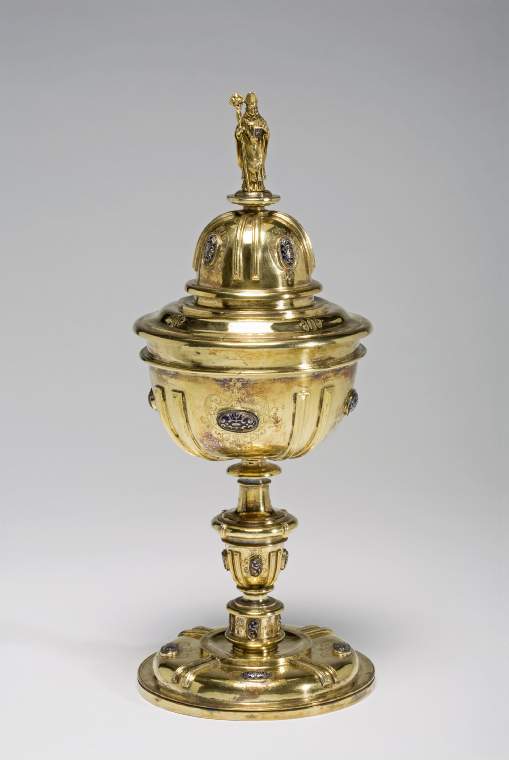 An image of ciborium and coversilver-gilt: set with enamelled silver bosses, the domed cover with finial of a bishopSilver-gilt; the stepped circular foot is engraved with four cartouches of scrolls each set with a silver boss enamelled in blue.  Between each boss is a pair of moulded ribs.  The stem comprises a series of spools and knops, set with two borders of bosses.  The top half of the U-shaped cup is undecorated.  Beneath an applied girdle the cup is set with four bosses between applied ribs to match the foot.  The circular cover stepped with a high central dome which is decorated with bosses and ribs to match the cup.  The top is surmounted by the cast figure of a bishop.