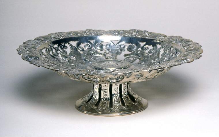 An image of Tazza. Standing dish. TC, silversmith, England, London. With heads of putti, sea monsters, snails, fruit, leaves and scrolls. Shallow circular dish with flat rim standing on a trumpet-shaped foot. The rim of the shaped circular dish is pierced, embossed and chased with alternating putti heads and fruit amongst scrolls. The dished area is pierced, embossed and chased with scrolling heads of sea monsters and snails alternating with winged putti heads. The slightly raised centre is pricked with a shield containing the initials 'I' over 'HI', around which are borders of punched circles and chased overlapping leaves and arches. The trumpet-shaped circular foot is pierced with vertical bands, and chased and punched with arches and dots. Silver, pierced, chased, punched and pricked, height, to rim, 8.5 cm, diameter, across rim, 28 cm, weight, whole, 555 g, circa 1616-circa 1617. Production Note: The form and design were probably influenced by Portuguese silver dishes.