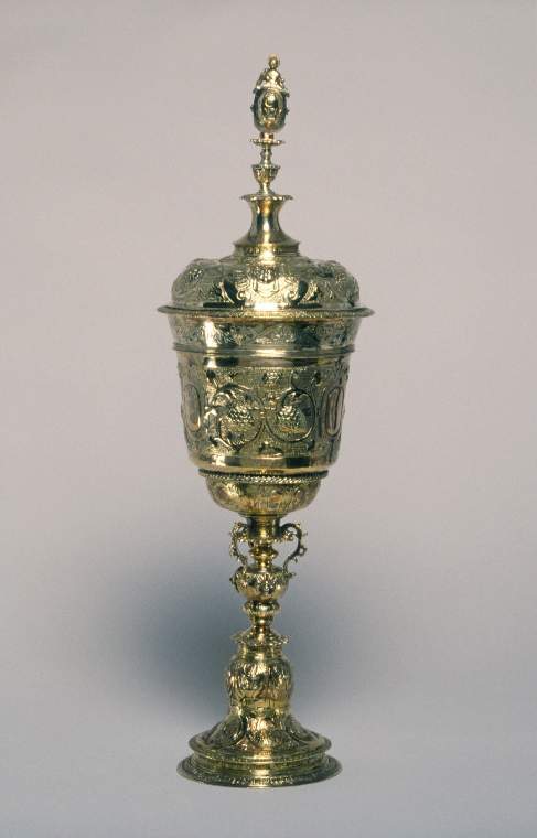 An image of standing cup and coversilver-gilt, u-shaped bowl on knopped stem above a bell-shaped foot, the domed cover with spool-shaped finial surmounted by a female figure, the body and cover embossed and chased.