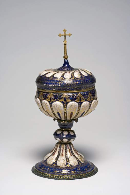An image of Cup and CoverCopper, enamelled and giltCopper, raised, enamelled in dark blue, dark green, dark red, and white enamels, and gilded; brass finial. The ciborium stands on a stepped and domed circular foot rising into a lobed stem supporting a separately made depressed globular knop with lobes above and below the equator, above which is a rounded calyx with everted petals on which the bowl rests. The deep rounded bowl is lobed round its lower part and has a gilt-copper rim. The domed cover has swirling gadroons on the top, and in the centre a finial in the form of a cross. The ground of both parts, including the interior of the bowl, and underside of the foot is dark blue. There are dark green borders round the foot and the base of the finial, and dark green petals on the calyx. The lobes on the foot, knop, bowl, and cover are white; and the ornament is executed in gold with red highlights on the lobed and gadrooned areas.Venice, ItalyCirca 1500