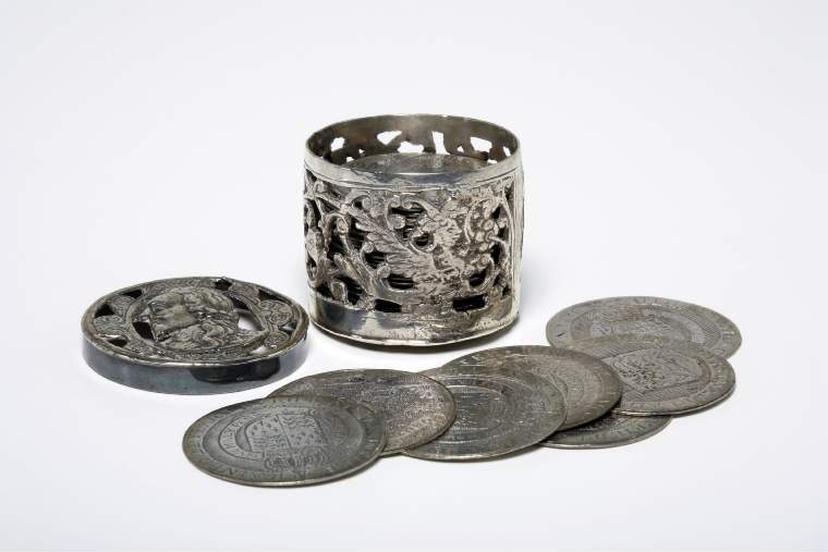 An image of Counter Box containing 32 countersCounter box. Silver, the box pierced, chased, and engraved, containing thirty-two engraved silver counters of English monarchs and their childrenCounter box. Silver, the box pierced, chased, and engraved; the counters engraved. The box is cylindrical with a detachable lid. The base is decorated with a bust of Queen Henrietta Maria in profile to left in low relief in an open oval medallion with pairs of inwards scrolls above and below, and S-scrolls on either side, with a cut away background. The top bears a bust of Charles I in profile to left in an open oval medallion surrounded by four paired scrolls with single scrolls between them, with a cut away background. The sides have two panels each containing pierced scrolling foliage and a monster. The counters are circular, and are engraved with monarchs and coats-of-arms respectively on the two sides: Prince Charles (d. 1130), Edward the Confessor, Willima I (2), William II, John, Edward I, Edward II, Edward III, Edward IV, Edward V, Edward VI, Henry I, Henry II, Henry III, Henry IV, Henry V, Henry VI (2), Henry VII, Henry VIII, Richard I, Richard II, Richard III, Mary I (2), Elizabeth I, Henry father of King James I and VI, James I and VI, Charles I, Elizabeth of Bohemia (his sister), Charles Louis Count of the Palatinate (her son)EnglishC.1640-50