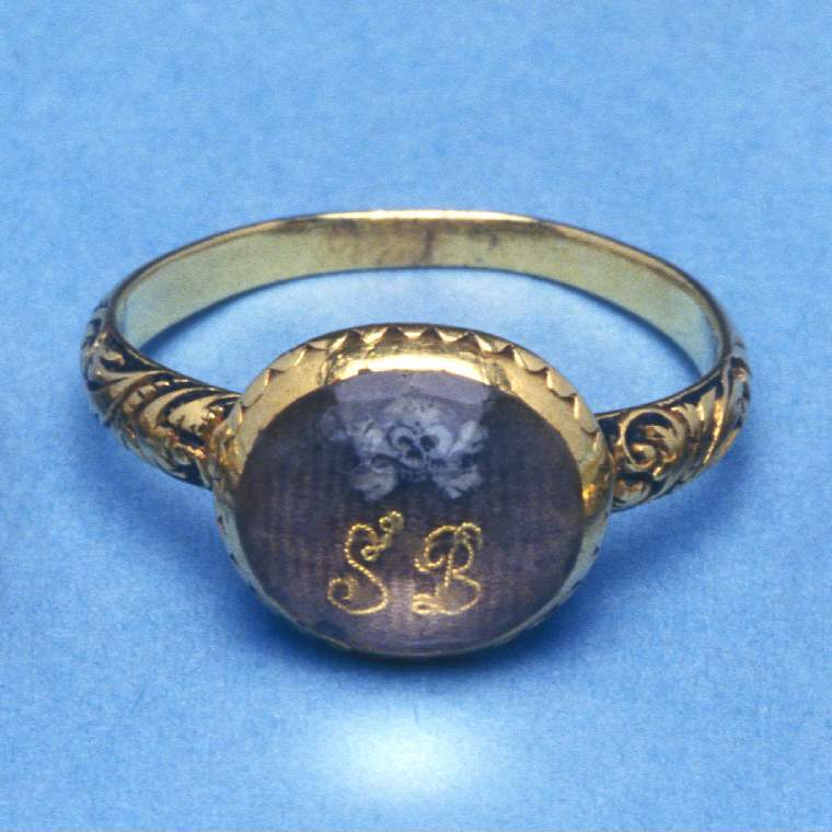 An image of Mourning ring. Gold, oval bezel with faceted glass (paste crystal?) covering the initials 'SB' in gold thread surmounted by a skull and crossbones, on a background of woven brown hair. Sides of bezel ornamented by fluting filled with black enamel and white dots. Shoulders of the plain gold hoop decorated with engraved foliate design reserved in black enamel.