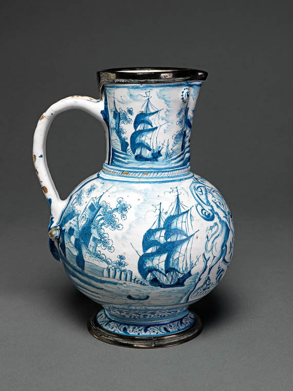 An image of JugEnglish delftwareTin-glazed earthenware painted in blue with the arms and motto of the Apothecaries' Company.Tin-glazed earthenware painted in blue. Bulbous body with pinched up lip, and on either side of it, a small embossment to stimulate an eye; grooved loop handled divided in the form of a fleur-de-lys at the lower end; standing on a low foot. Both rim and foot mounted in silver. Decrated on the front with the full arms of the Apothecaries' Companoy with motto: 'OPIFER:QVE:PER: ORBEM:DICOR'; at the sides and on the neck, sea-shore scenes with buildings and sailing-ships. Two narrow borders of leafy scrollwork encircle the foot. Belowthe handle are the initials and date 'E.V/1650'. The mounts bear the London hallmark for 1844.Southwark, LondonEnglandC.1650