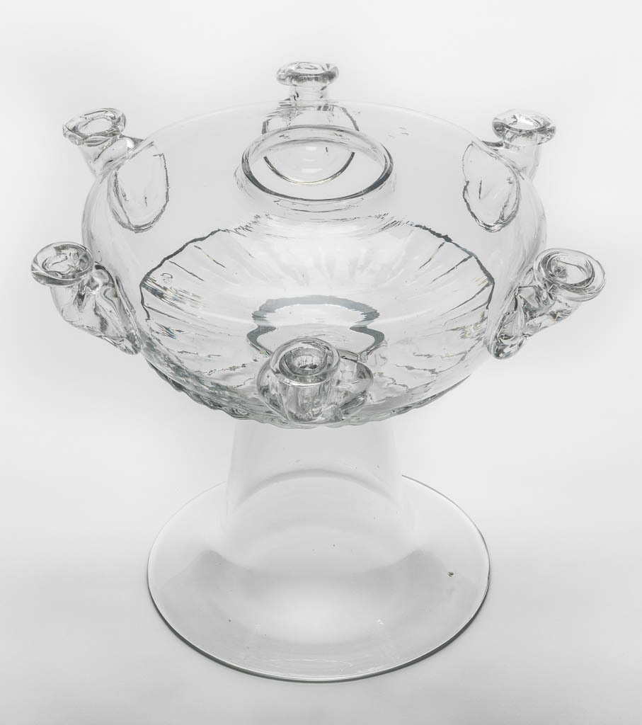 An image of Glass Lamp. Unknown English glassmaker. The depressed globular bowl of the lamp is blown-moulded with gadrooning round the lower part, six applied nozzles for wicks at regular intervals on the sides, and a circular hole in the centre. It is joined to a tall, plain inverted trumpet-shaped foot. Clear lead glass, lamp bowl blown-moulded, nozzles applied decoration, height, whole, 22.0 cm, diameter, foot, 15.7 cm, circa 1700-1725. Baroque. Queen Anne, George I.