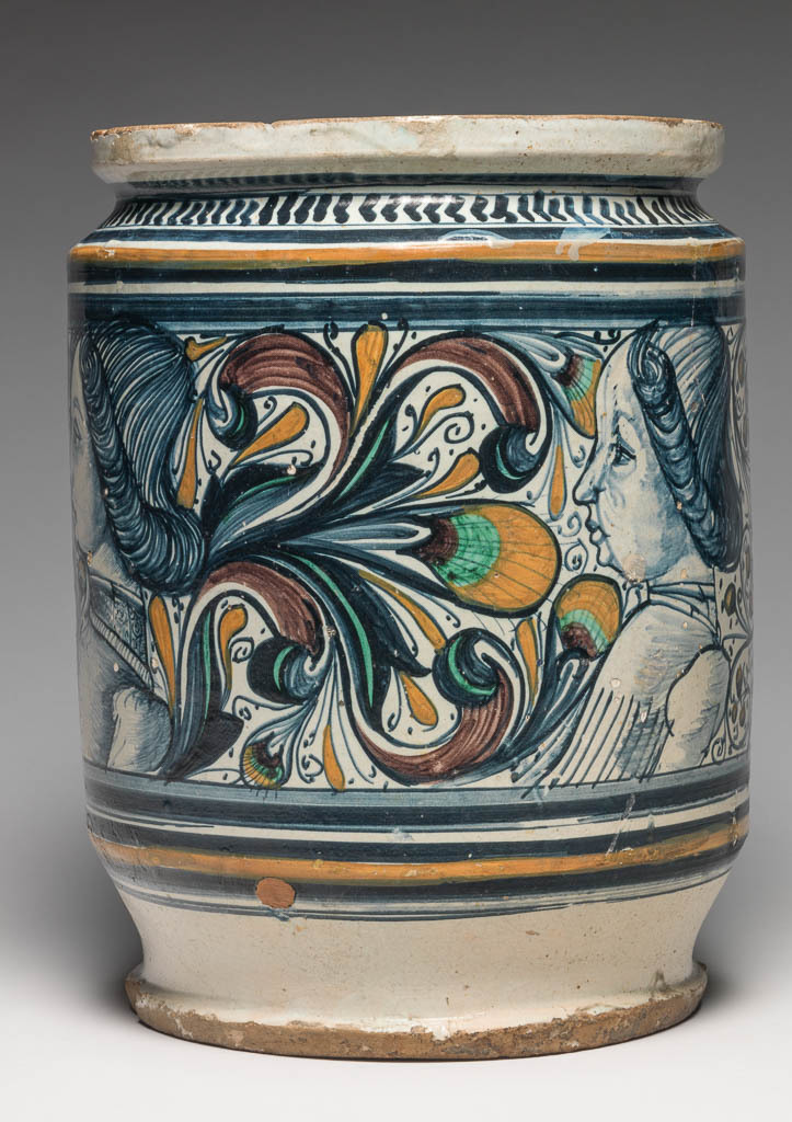 An image of Maiolica. Albarello/Pharmacy Jar. Unidentified potter, The Marches, Pesaro. Formerly attributed to Faenza. Broad albarello form with straight sides. Round the middle are four busts of boys wearing contemporary costume face in profile to left. One is flanked by gothic foliage and peacock's feather eye motifs, the others by scrolling foliage bearing parti-coloured fruits against a dotted background. Above and below there is a wide blue band and a dark yellow band between two blue; and on the shoulder, a row of blue chevrons with a blue band above. Buff earthenware, thrown, tin-glazed on the exterior and very thinly on the interior; base unglazed. Painted in dark blue, green, dark yellow, and manganese-purple, height, whole, 26.8 cm, diameter, rim, 18.8 cm, diameter, base, 17.2 cm, diameter, whole, 20.3 cm, circa 1470-1490. Renaissance.