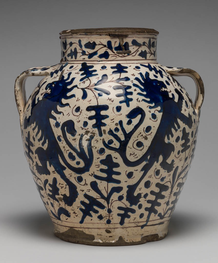 An image of Maiolica. Pharmacy or storage jar. Unknown maker, Florence. Both sides are decorated with two lions rampant addorsed, each holding a spray of oak leaves in its raised paw, surrounded by sprays of oak leaves with dots in the background. Round the lower part of the body there are two horizontal manganese bands. On the neck, between pairs of horizontal manganese bands, there are oak leaves on a wavy stem. On each handle, and continuing down the side of the jar beneath it, is a fern frond or stalk with twigs branching off on each side and terminating in small round berries. Buff earthenware, tin-glazed creamy-white on the interior and exterior; the rim and base unglazed. Painted in bright, extremely shiny relief-blue with manganese outlines. Height, whole, 25.8 cm, diameter, mouth, 12.4 cm, diameter, base, 14.5 cm, width, across handles, 23.0 cm, circa 1420-1450. Renaissance.