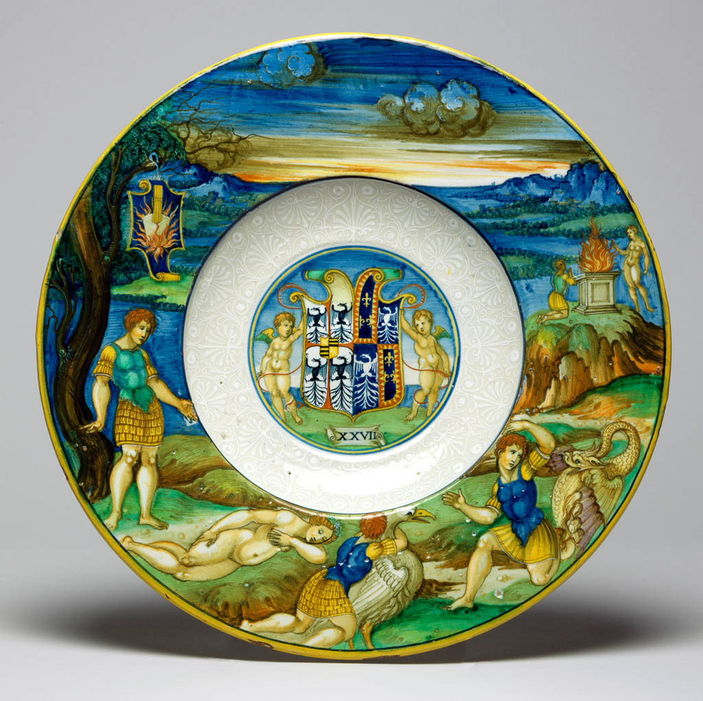 An image of Maiolica. Dish from the Isabella d'Este Service. Nicola di Gabriele Sbraghe da Urbino (Italian, d.1537/38). Shape 56. circular with broad, almost flat rim, and deep well; the underside of the rim moulded with three bands of reeding. In the well, within narrow blue and yellow concentric circles, two putti support a shield charged with the arms of Gonzaga impaling Este: argent, a cross patty gules cantoned by four eagles displayed sable (Gonzaga modern granted 1433), an escutcheon overall, quarterly, 1, 4, gules a lion rampant argent (Bohemia), 2, 3, barry of six or and sable (Gonzaga ancient) impaling Este, quarterly, 1, 4, azure three fleurs-de-lis or a bordure indented gules, 2, 3, azure an eagle displayed argent (gules shown orange throughout). Below on a scroll is the impresa `XXVII'. The sides are decorated in bianco sopra bianco with palmettes and scrolls, and a blue line encircles the shoulder. The rim is decorated with Peleus and Thetis. Peleus stands beneath a tree, gazing at Thetis, who sleeps nude on the ground in front of him. Further to the right she is transformed into a swan-like bird as he embraces her, and then into a dragon from which he turns away in horror. Higher up on the right, a goddess appears to Peleus who kneels before an altar. In the distance there is a mountainous landscape and sky. Suspended from the tree there is a shield of testa di cavallo form charged with the impresa of gold rods in a crucible surrounded by flames in a blue ground. Earthenware, tin-glazed overall; the reverse pale beige and speckled. Painted in blue, green, yellow, orange, stone, brown, manganese-purple, black, and white high-temperature (metallic oxide) colours, height, whole, 3.9 cm, diameter, rim, 30.2 cm, circa 1524-1525. Renaissance. Notes: The figures were derived from a woodcut illustration in Giovanni dei Bonsignori's Ovidio methamorphoseos vulgare, Venice (Giovanni Rosso da Vercelli for Lucantonio Giunta), 1497 or a later edition.