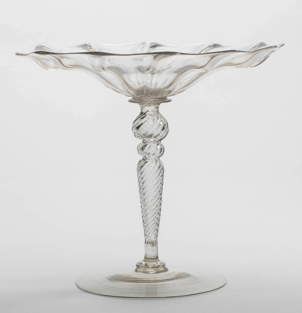 An image of Glass/Tazza. Unknown, glasshouse, The Veneto. Open flower like bowl with wavy edge, and ribbing on the underside. Inverted baluster stem with two knops, all ribbed spirally. Plain foot. Lead-glass, moulded, with ribbed decoration, height, whole, 15.7 cm, diameter, whole, 7.0 in, circa 1575-1625. Renaissance.