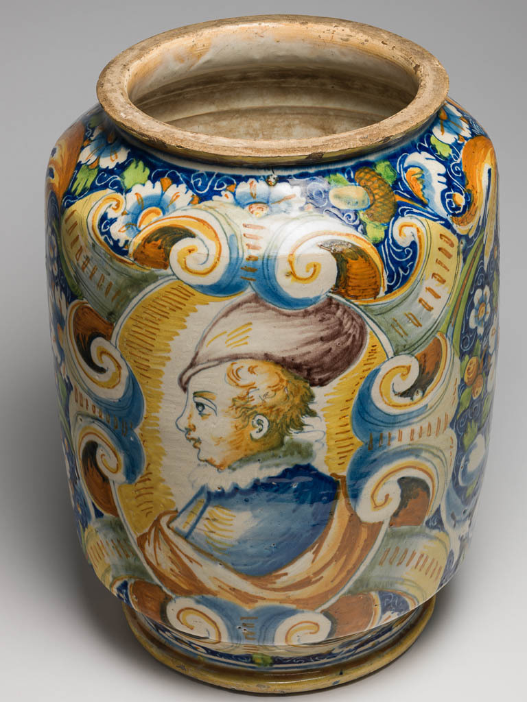 An image of Maiolica. Pharmacy jar/storage jar. Domenego da Venezia, workshop, The Veneto. On the front is a rectangular label inscribed `Mostarda FA' in blue gothic script, and framed by scrolls with a face at bottom centre. On the other side, within an oval scrolled frame, is a bust of a young man in profile to left, wearing a doublet, ruff and a soft rounded hat. The rest of the main field is decorated with stylised flowers, fruits, acorns and scrolling foliage, reserved in a blue ground incised with coiling tendrils. Earthenware, thrown, tin-glazed on the exterior and interior; rim and base unglazed, painted in polychrome, height, whole, 39.5 cm, diameter, rim, 21.5 cm, diameter, base, 22.0 cm, width, widest part, 28.9 cm, circa 1560-1570. Renaissance.