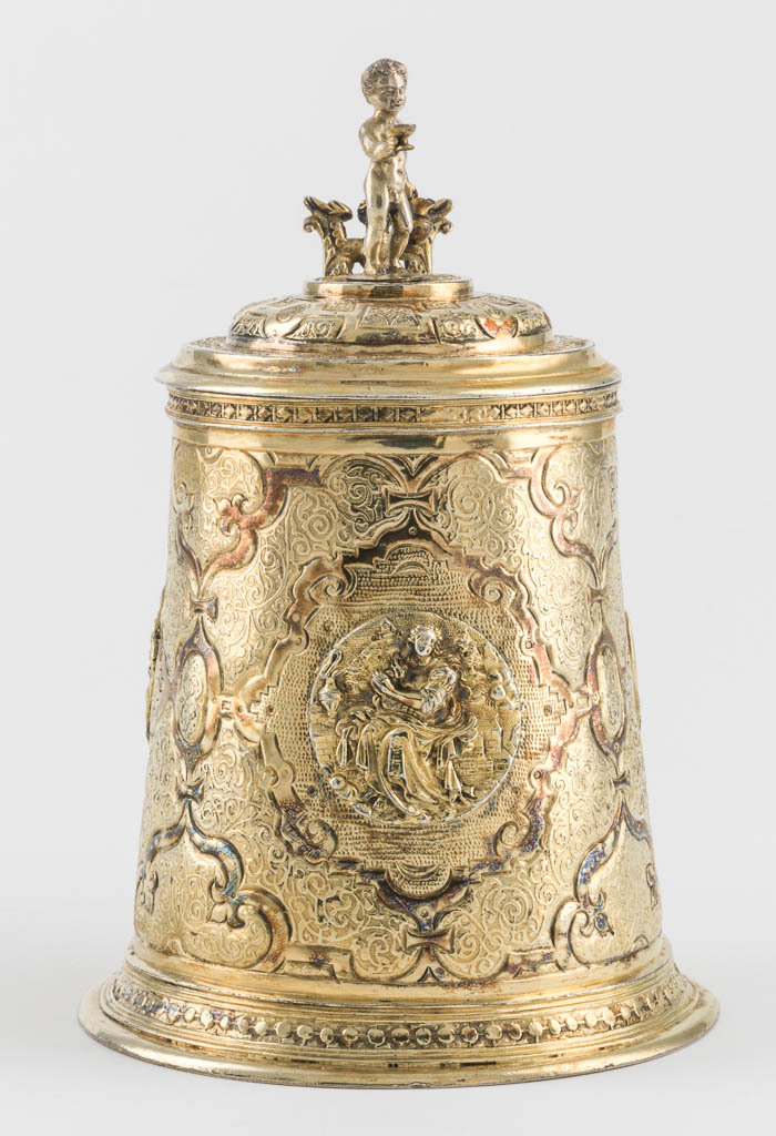 An image of Tankard. SA, silversmith, Augsburg. Flötner, Peter, after (German, c.1485-1546). The sloping cylindrical sides are embossed and chased with an elaborate pattern of straps containing etched Arabesques and three applied cast relief medallions of Charity, Hope and Temperance. The similarly decorated stepped cover is sumounted by a figure of a young Bacchus and has a thumb-piece in the form of a double tailed mermaid. The spreading circular foot and the rim of the neck are decorated with a border of flattened beads. The D-shaped handle is engraved with a border of Arabesques and has an applied acanthus leaf at the top. Silver whole, gold surface (silver-gilt, embossed, chased and etched), height, to top of finial, 17 cm, diameter, base, 11.3 cm, weight, whole, 480 g, circa 1570-1600. Renaissance. Notes: The young Bacchus finial is almost certainly a replacement. The gilding is later. The plaques of Charity, Hope, and Temperance are after plaquettes by a follower of Peter Flötner, after rectangular plaques by Flötner.