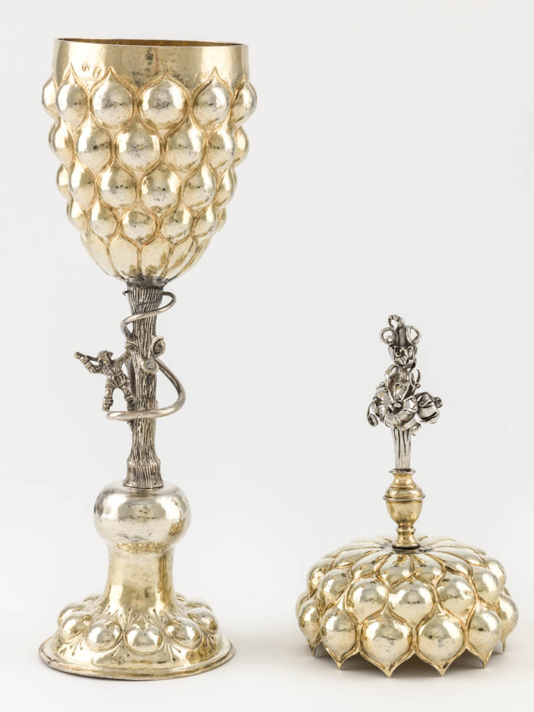 An image of Cup and cover/pineapple cup. Fischer, Franz (silversmith, Nüremberg, surname is also spelt Vischer). Lobed ovoid body and cover supported by a stem in the shape of a tree trunk with figure of a woodman. Silver-gilt; the spreading circular silver-gilt foot is embossed with a ring of lobes round a central dome, which supports the silver stem in the shape of a tree trunk encircled by a creeper, and the figure of an ax-wielding woodman. The ovoid silver-gilt body and interlocking cover are embossed with lobes. The silver finial is formed as a bunch of flowers. Silver and gold, raised and embossed body and foot, cast stem and woodcutter, height, overall, 33.3 cm, diameter, overall, 8 cm, weight, whole, 281 g, 1600-1653. Mannerist.