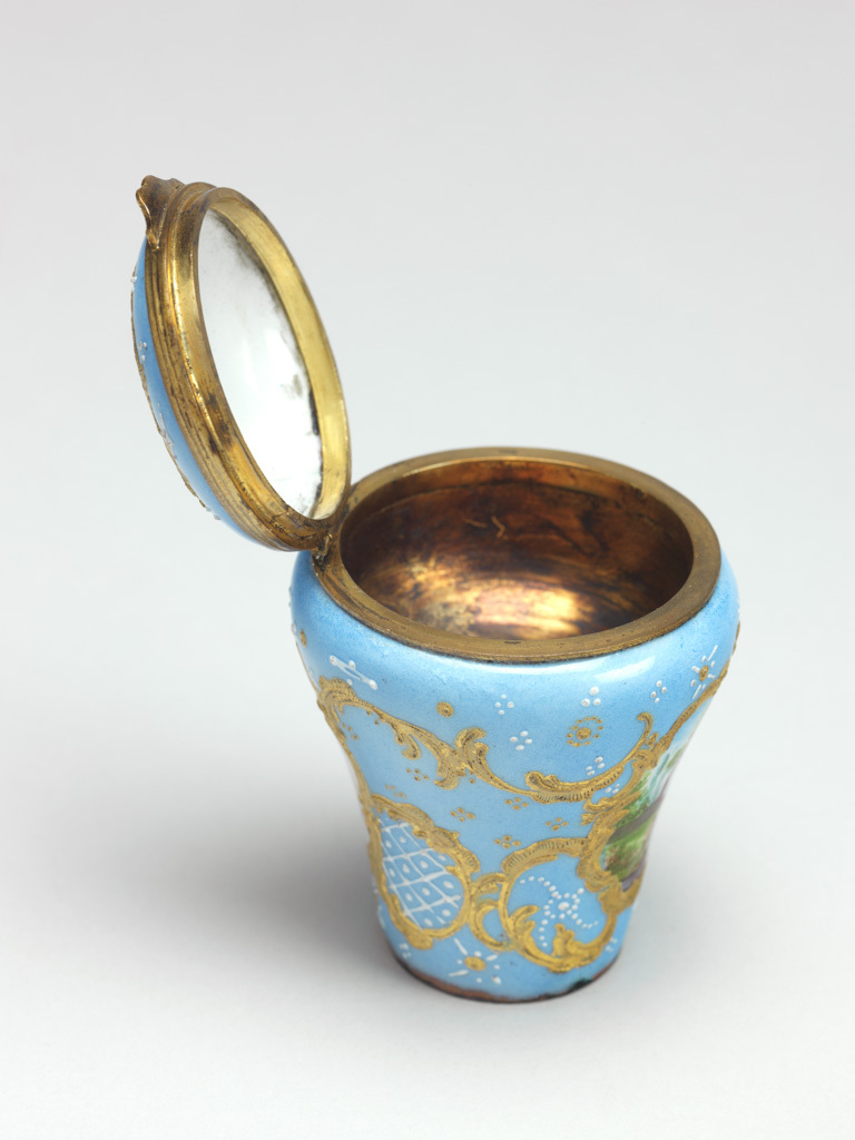 An image of Cane Handle/Snuff Box. Objets de vertu. Unknown maker, Staffordshire. Baluster-shaped handle for a walking cane set with a hemi-spherical gilt metal snuff box with a hinged lid. On two sides and the lid are painted miniatures within rococo raised gilt borders. The pale blue ground decorated with white diaper-work and geometric patterns of gold and white dots. The rim of the lid and box have gilt metal mounts and thumbpiece. The miniature on the lid depicts a Classical ruin in a pastoral scene; the miniature on the front depicts a man and cow in a rural landscape with a church tower and cottage in the background, and the miniature on the back depicts a spray of flowers on a white ground. Copper, enamelled in polychrome, with gilt metal mounts, height, whole, 5.5 cm, diameter, whole, 4.5 cm, circa 1750-1775.