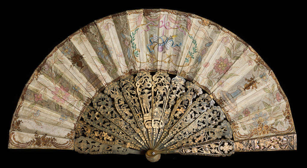 An image of Marriage Fan/Folding Fan. Double leaf of silk, painted in bodycolour, gold and silver, and decorated with gold tambour work and sequins. Sticks and guards of horn (12+2), shaped, pierced, silvered, gilt and set with clear pastes brilliants; guards also backed by irridescent shell. Rivet set with clear pastes. Length, guards, 25.2 cm, circa 1760.