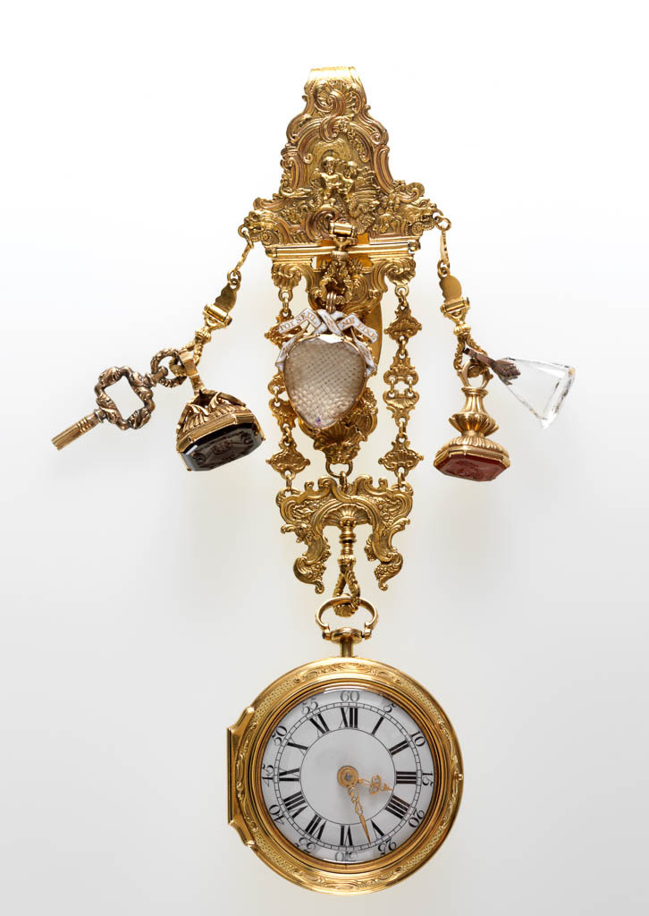 An image of Watch and chatelaine. Goujon, Stephen (case maker, London). Moser, George Michael (gold chaser, case, British, 1704/1706-1783, London). Webster, William (watchmaker, active 1734-1766, London). The gold watch case embossed and chased on the front with Venus presenting Arms to Aeneas watched by a river god and surrounded by Rococo scrolls. Chatelaine is cast with rococo designs, back of large central plaque with incused 'H' under a vertical line of five chevrons. Side drops, left hand with key and seal, cut with coat of arms, right hand with two seals, one cornelian cut with head of a philosopher, and one rock crystal cut with a head of a bearded man, possibly Socrates'. Centre drop at top with heart-shaped locket containing woven hair under under glass, above which is a white enamelled gold ribbon with gold lettering 'TOT SEUL ME FIXE.'. Case, gold, with chased decoration; chatelaine, cast and chased, height, whole, 14.1 cm, 1761-1762. Rococo.