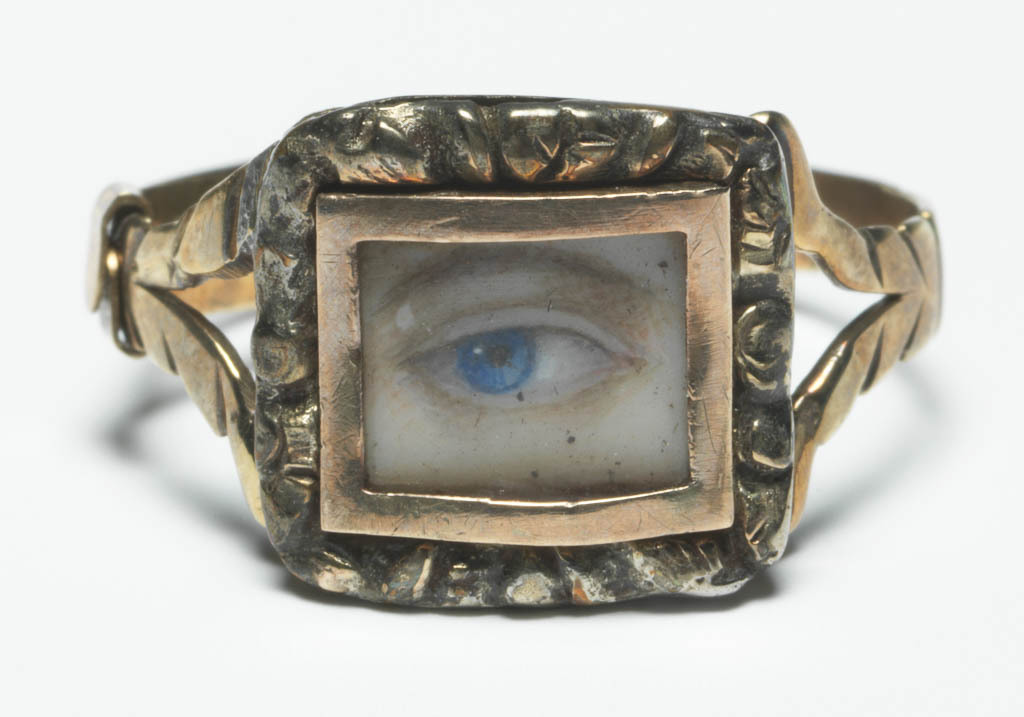 An image of Jewellery/Ring. The ring has a thin gold hoop dividing into two branches at the shoulders, and a rectangular silver bezel with cast and chased border, in which is set a rectangular, plain gold frame enclosing under glass a miniature of a brown eyebrow and an eye with a blue iris. Gold, silver, glass and painted miniature on ivory, height, whole, 1.9 cm, circa 1790-1800. English.