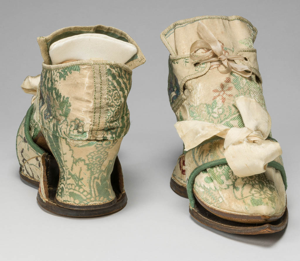 An image of Textiles/Shoes and pattens. Brocaded silk, cream background, multicoloured floral design. Shoes lined with coarse linen, tongues with green sarcenet, bound green braid. Pattens lined scarlet flannel, bound green braid, cream corded silk bows, leather soles and heels. Length, whole, 8.25 in, circa 1700-1749. English.