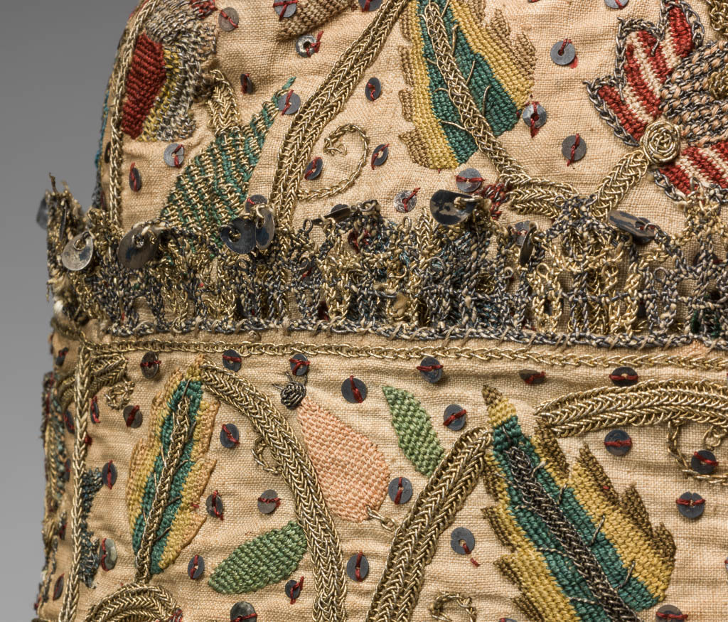 An image of Textiles. Man's cap. Linen, embroidered with polychrome silks, silver and silver-gilt threads. The brim is edged with metal thread bobbin lace. The cap comprises four joined panels each decorated with coiling stems carrying leaves, flower sprays, insects and birds. Height, whole, 11¼ in, width, whole, 9 in, circa 1575-circa 1599. English. Elizabethan.
