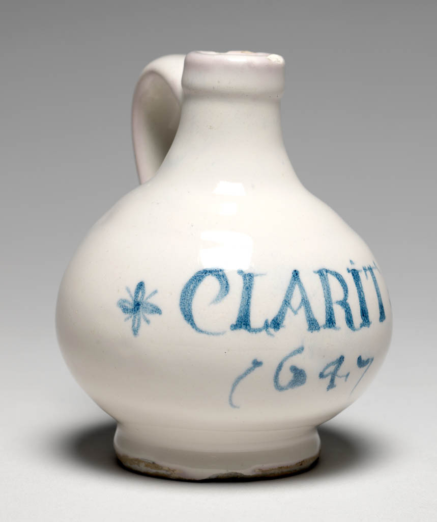 An image of English Delftware. Wine bottle/wine jug. Probably made in Southwark, or possibly in Brislington. The jug has a low foot, a bulbous body, short slender neck with a projecting band round the top, and loop handle which is rounded on the exterior. On the front is the inscription CLARIT/ 1647 with a star on each side. Buff earthenware, thrown, with slightly concave underside to the foot, and a groove round its edge, and an applied handle, tin-glazed, and painted in blue; the glaze has been wiped off most of the base, height, whole, 11.6 cm, dated 1647. English.