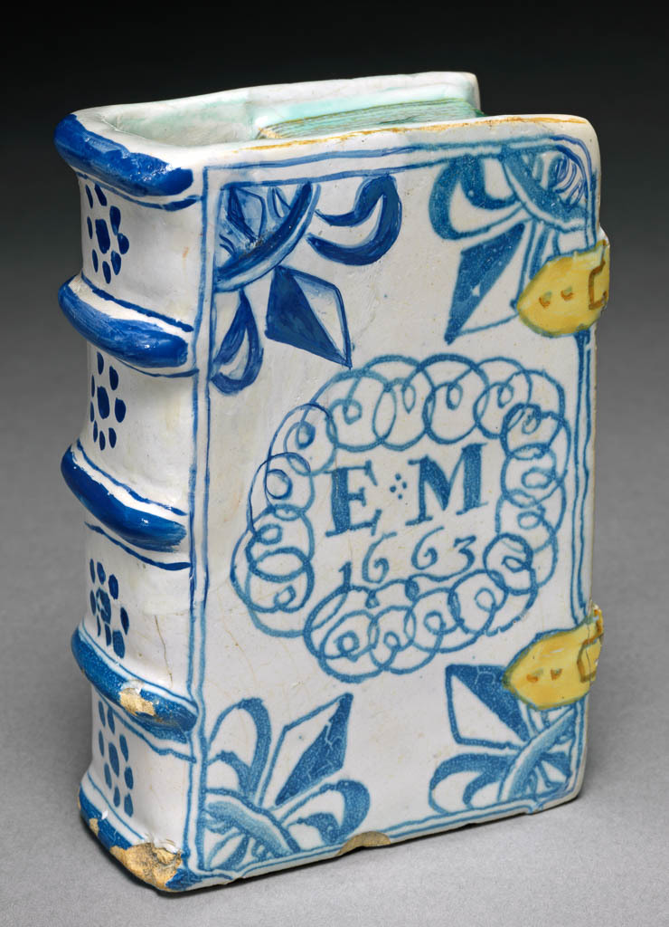 An image of English Delftware. Hand Warmer in the Shape of a Book. Unidentified London pottery, or possibly Brislington Pottery. White earthenware, tin-glazed, and painted in blue, turquoise-green, yellow and ochre. Formed as a book with two false clasps, the hand warmer has two compartments linked by a hole, and having a large approximately rectangular aperture in the top near the spine, and a small hole in the back top left corner. Both sides are painted with the initials and date 'EM/1663' within an oval loopy frame, and have a fleur-de-lys in each corner. The spine is painted with four horizontal stripes alternating with dotted flowerheads, and the fore-edge is painted in turquoise with slightly darker stripes to indicate the pages. Height, whole, 11.3 cm, width, whole, 7.9 cm, depth, whole, 4 cm, dated 1663.