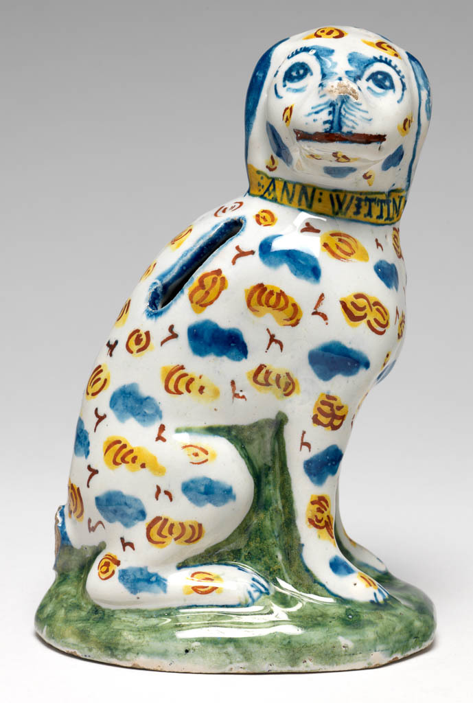 An image of English Delftware. Money Box in the shape of a Dog. Brislington Pottery, Somerset. It wears a yellow collar inscribed 'ANN WITTIN 1717' in blue. The unglazed base is inscribed in blue, 'Ann Wittin/was born ye 14 of/November 1717 J (?) W'. Buff earthenware, moulded with pierced slot to take money, tin-glazed except for the base, and painted in blue, green, yellow, and red high-temperature (metallic oxide) colours, height, whole, 14.9 cm, dated 1717.