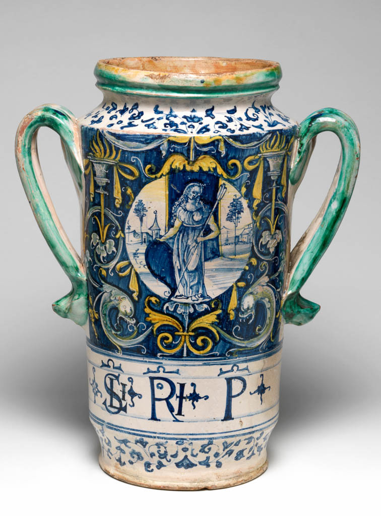 An image of Tin-glazed Earthenware / Two-handled pharmacy jar or drug jar. On the front, a woman in a landscape surrounded by grotesques. Red earthenware, the interior lead-glazed; the exterior tin-glazed, but applied thinly on the back and appearing pinkish-grey in places. Painted in blue, green, and yellow. Maiolica, height, whole, 37.0 cm, diameter, rim, 15.8 cm, diameter, base, 15.5 cm, width, whole, 30.1 cm, circa 1510-1530. Renaissance. Italy, Tuscany, Siena (perhaps).