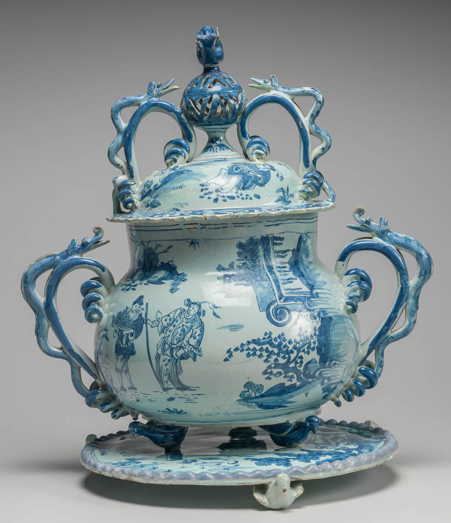 An image of English Delftware. Posset pot with cover and stand (see C.1504 & B-1928). Brislington Pottery, Somerset (possibly). The pot is bulbous, with wide neck and two handles branching into corkscrew coils and decorated with applied serpents; between them, on one side, is a sucking spout. On the cover, which is domed, is a knob in the form of an openwork globe with a bird perched upon it, flanked by handles similar to those on the pot. The pot and the circular stand each rest on three feet in the shape of small birds. The pot and the cover are painted with Chinese figures in landscapes, in the manner of late Ming porcelain; under the base the initials and date 'C T A 1685'. On the stand there is a man and woman in European dress drinking (perhaps intended as Lot and his daughters), with sun, moon and stars in the sky and the same initials and date '1686'. Buff earthenware, the body thrown, with applied handles, spout, feet, pierced knob and bird finial, covered with very pale turquoise-green tin-glaze, painted in blue. Height, whole, 31.1 cm, width, whole, 31.0 cm, dated 1685 (stand 1686). Baroque. Late Ming (manner of). Production Note: Previously uncertain whether the posset pot was Lambeth or Bristol. See Garner, 1972, in documentation, for attribution to Lambeth. See Archer, 2007 for attribution possibly to Brislington. The stand is catalogued separately because it has unrelated decoration, and may not originally have been with this posset pot.