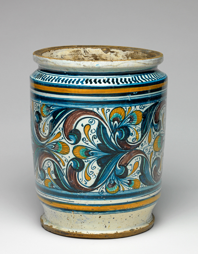 An image of Maiolica. Pharmacy jar/albarello. Unidentified Pesaro potter, The Marches. Broad albarello with straight sides. On the front, in a contour panel with three lobes on each side, are profile busts of a man and woman wearing contemporary costume, facing inwards towards a vase of pinks, with groups of three dots in the background. The rest of the main field is decorated with gothic foliage and peacock's feather eye motifs. Above and below there is a wide blue band and a dark yellow band between two blue; on the shoulder, a row of blue chevrons with a horizontal blue band above. `4S758' is incised into the base. Buff earthenware, tin-glazed on the exterior and more thinly on the interior; base and outer edge of base unglazed. Painted in dark blue, green, dark yellow, and manganese-purple. Height, whole, 26.9 cm, diameter, rim, 20.0 cm, diameter, base, 17.3 cm, diameter, whole, 21.2 cm, circa 1470-1480. Renaissance.