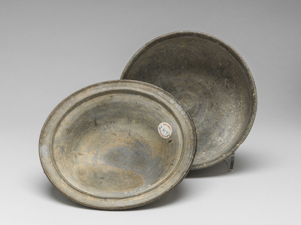 An image of Vessel/Bowl. An almost hemispherical bowl on a low thick foot-rim of grey earthenware with a bowl shaped lid with a small circular knop which could serve for a foot. Impressed with a pattern of radial strokes and small circles with a centre dot, with traces of grey-black glaze. 400-699 AD. Early Silla Period. Korean.