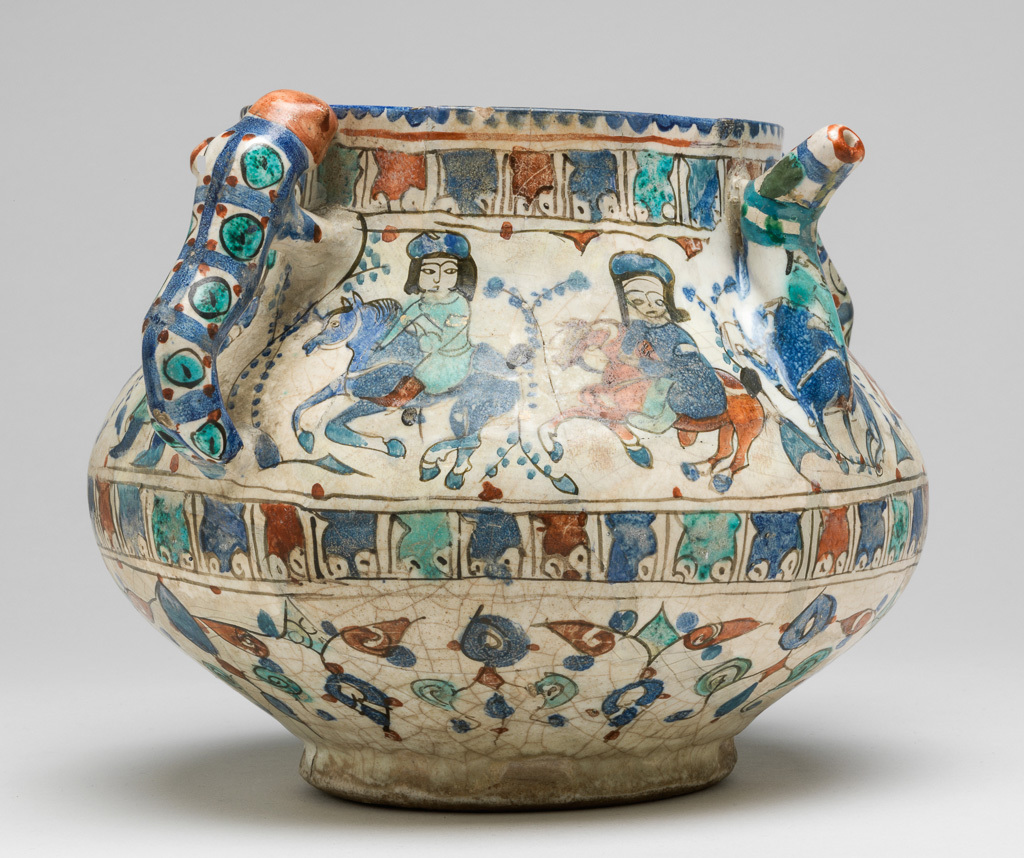 An image of Posset pot/spouted jar. Islamic pottery, Minai. Unknown, potter, probably Kashan. Shape: bulbous body with a cylindrical neck and plain rim, sitting on a low foot ring. Opposing cheetah handles and angular spouts are attached to the upper body. Exterior: the rim is painted with a blue dentilated pattern. On the upper body, a frieze of mounted horsemen and two standing figures each separated by feather-like foliage, are identically dressed in hats, tunics, and leggings, occasionally wearing golf leaf arm bands. This frieze is delineated above and below by narrow bands of Arabic script, possibly pseudo epigraphic. On the lower body, a frieze of alternately transposed triangular floral garlands is painted. All these friezes are outlined in black and coloured with blue, turquoise, red and black. Two lion shaped handles, with red heads and bodies decorated with blue stripes and turquoise and red dots, are joined to the upper body. White glaze runs unevenly over the lower body and in places onto the foot ring. Interior: blue paint continues on the rim, while on the neck black Arabic script (cursive) is painted. Remainder is undecorated except for a covering of white glaze. Fritware, wheel thrown body and moulded handles and spout, coated in an opaque white, slightly crazed glaze; decorated with blue and turquoise in-glaze painting, black and red overglaze painting and gilding. Height, whole, 14.5 cm, width, whole, 17.4 cm, diameter, rim, 11.3 cm, diameter, base, 8.9 cm, weight, whole, 708 g, circa 1180-1219. Seljuk Period.