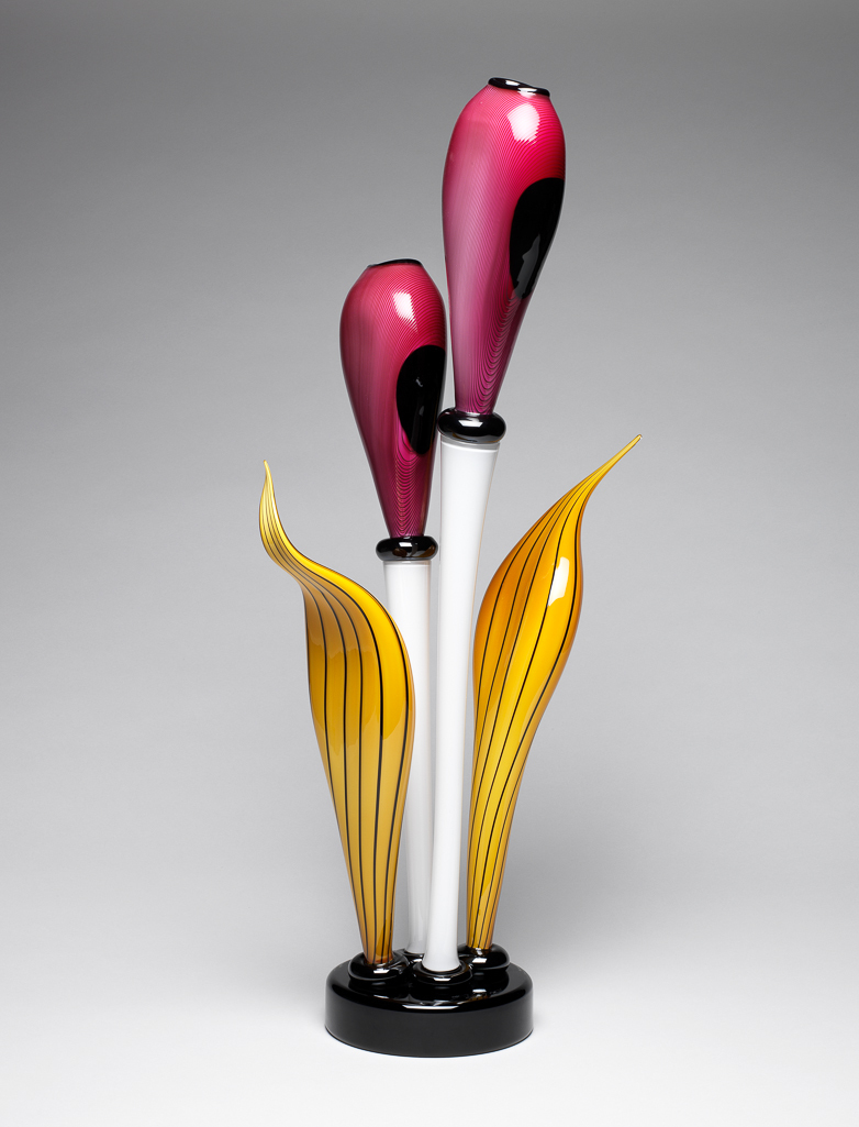 An image of Contemporary Craft. Studio Glass. Flower container. Narcissus III. Crooks, Robert S (British, b. 1965). The flower containers are two flattened ovoid buds with an opening at the top, supported by tubular stalks, flanked by two long pointed leaves, one with a kink near to its tip, rising from four anulets on top of a circular, straight-sided, hollow base. The pink buds are striped in dark pink, and each has two oval black spots, a black band round the mouth, and a black anulet at the top of the white stem. One stem is taller than the other. The dark yellow leaves have black stripes. The base, and the four anulets round the base of the stems are black. Pink, dark pink, dark yellow, white, and black hand-blown glass. height, whole, 66.4 cm, width, whole, 33.5 cm, diameter, base, 14.7 cm, 2000. Gift of Nicholas and Judith Goodison through the National Art Collections Fund.