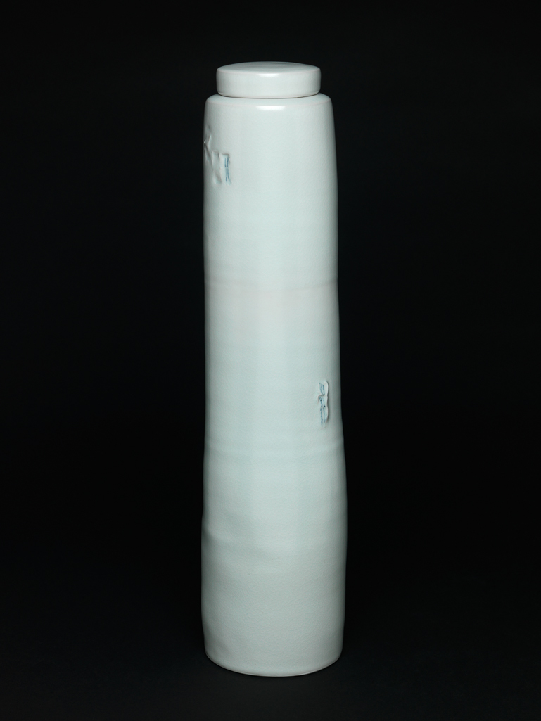 An image of Studio Ceramics. Contemporary Craft. Tall Lidded Jar. Waal, Edmund de (British, b.1964). Porcelain, thrown, and impressed in two places with seal marks coloured blue under pale celadon glaze. Cylindrical with uneven, slightly tapering sides and visible throwing marks, narrow shoulder, and short unglazed neck. The base has a glazed central circular depression surrounded by a concave area, and a flat unglazed edge which has been scored with criss-cross lines. The sides are decorated in two places with impressed 'seal marks, touched with blue: a group of three near the top, and one further to the right about half way down The circular cover has straight sides and very slightly convex top. Its interior is unglazed except for a circle in the centre. A black felt circle has been applied to the outer edge of the base. Height, whole, 50 cm, diameter, whole, 12.2 cm, diameter, cover, 8.5 cm, 1999. Gift of Nicholas and Judith Goodison through the National Art Collections Fund.