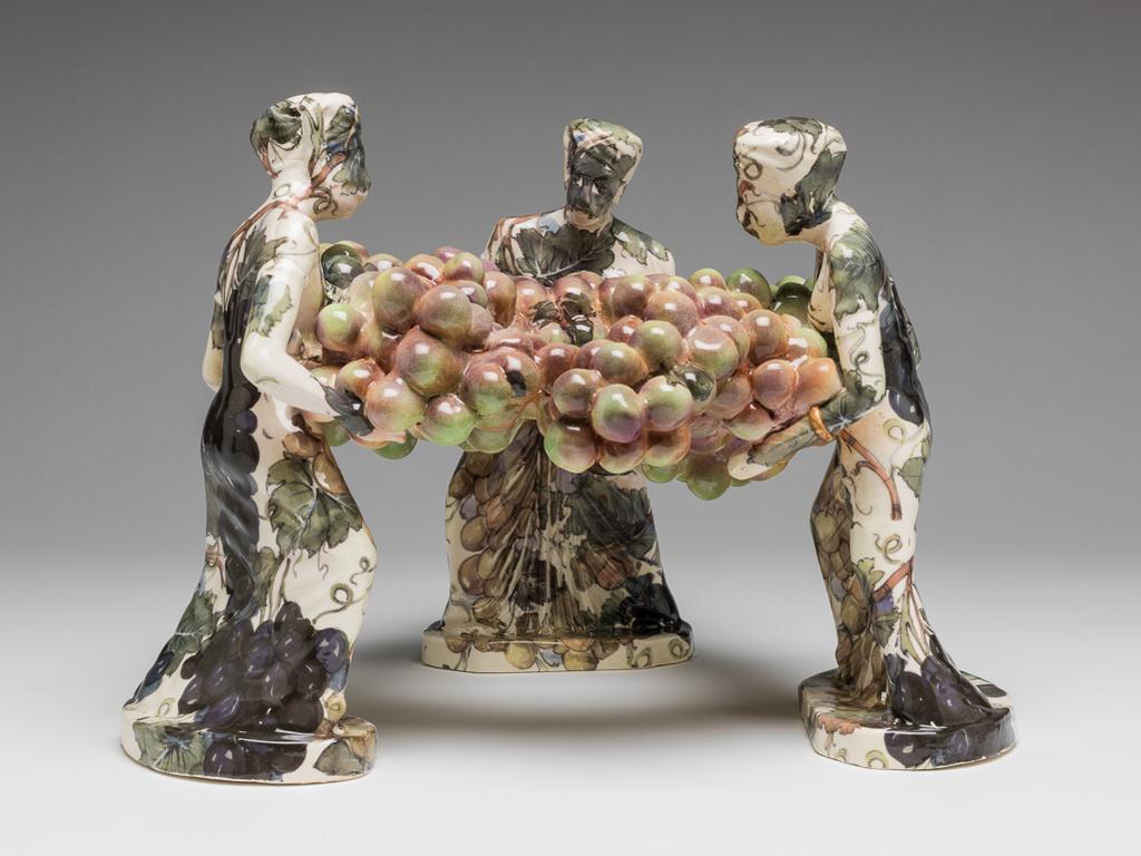 An image of Studio Ceramics. Banana Dish. McNicoll, Carol (British, b. 1943). Three turbaned figures in long pleated gowns standing on oval bases, holding between them a garland of grapes. The figures are decorated all over with fruiting vine in shades of greyish-green, purple, pink, and pale brown, and have narrow gold bracelets. The garland of grapes is painted underlgaze in pale green, and pinkish-purple, with transferred leaves here and there. The underside of each base is pale green. The base of one figure is titled 'BANANA/DISH' in black. Cream earthenware, slip-cast, painted underglaze in pale green and pinkish-purple, and decorated with open stock French slide on screen-printed ?transfers. Height, whole, 23.4 cm, width, whole, 32.5 cm, 1999. Gift of Nicholas and Judith Goodison through the National Art Collections Fund.