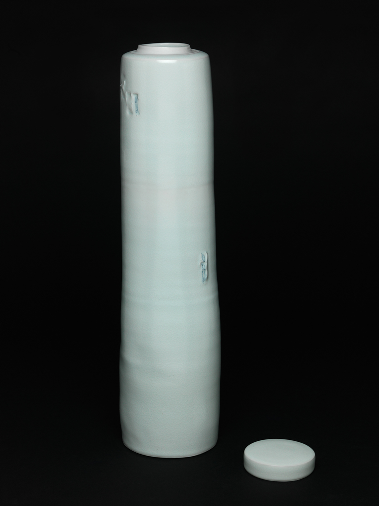 An image of Studio Ceramics. Contemporary Craft. Tall Lidded Jar. Waal, Edmund de (British, b.1964). Porcelain, thrown, and impressed in two places with seal marks coloured blue under pale celadon glaze. Cylindrical with uneven, slightly tapering sides and visible throwing marks, narrow shoulder, and short unglazed neck. The base has a glazed central circular depression surrounded by a concave area, and a flat unglazed edge which has been scored with criss-cross lines. The sides are decorated in two places with impressed 'seal marks, touched with blue: a group of three near the top, and one further to the right about half way down The circular cover has straight sides and very slightly convex top. Its interior is unglazed except for a circle in the centre. A black felt circle has been applied to the outer edge of the base. Height, whole, 50 cm, diameter, whole, 12.2 cm, diameter, cover, 8.5 cm, 1999. Gift of Nicholas and Judith Goodison through the National Art Collections Fund.