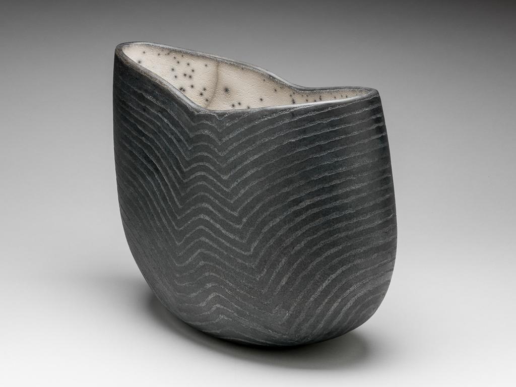 An image of Contemporary Craft. Studio Ceramics. Vessel. Roberts, David (British, b.1947). Of elongated oval plan, with curved slides, rising higher at one end than the other, so that the rim slopes downwards in a gentle curve on that side, and then upwards slightly to the other side. The exterior is very dark grey, and is decorated with horizontal lines which echo the curvature of the rim, and create an optical illusion that the vessel has an inverted V-shaped channel on each side. The interior is greyish-white slip with deliberate grey cracks and numerous dark grey spots. Stoneware, hand-built, and raku glazed, height, whole, 28.5 cm, length, whole, 35.0 cm, 1998. Gift of Nicholas and Judith Goodison through the National Art Collections Fund.