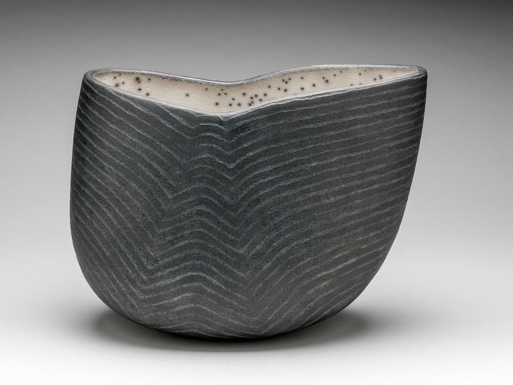 An image of Contemporary Craft. Studio Ceramics. Vessel. Roberts, David (British, b.1947). Of elongated oval plan, with curved slides, rising higher at one end than the other, so that the rim slopes downwards in a gentle curve on that side, and then upwards slightly to the other side. The exterior is very dark grey, and is decorated with horizontal lines which echo the curvature of the rim, and create an optical illusion that the vessel has an inverted V-shaped channel on each side. The interior is greyish-white slip with deliberate grey cracks and numerous dark grey spots. Stoneware, hand-built, and raku glazed, height, whole, 28.5 cm, length, whole, 35.0 cm, 1998. Gift of Nicholas and Judith Goodison through the National Art Collections Fund.