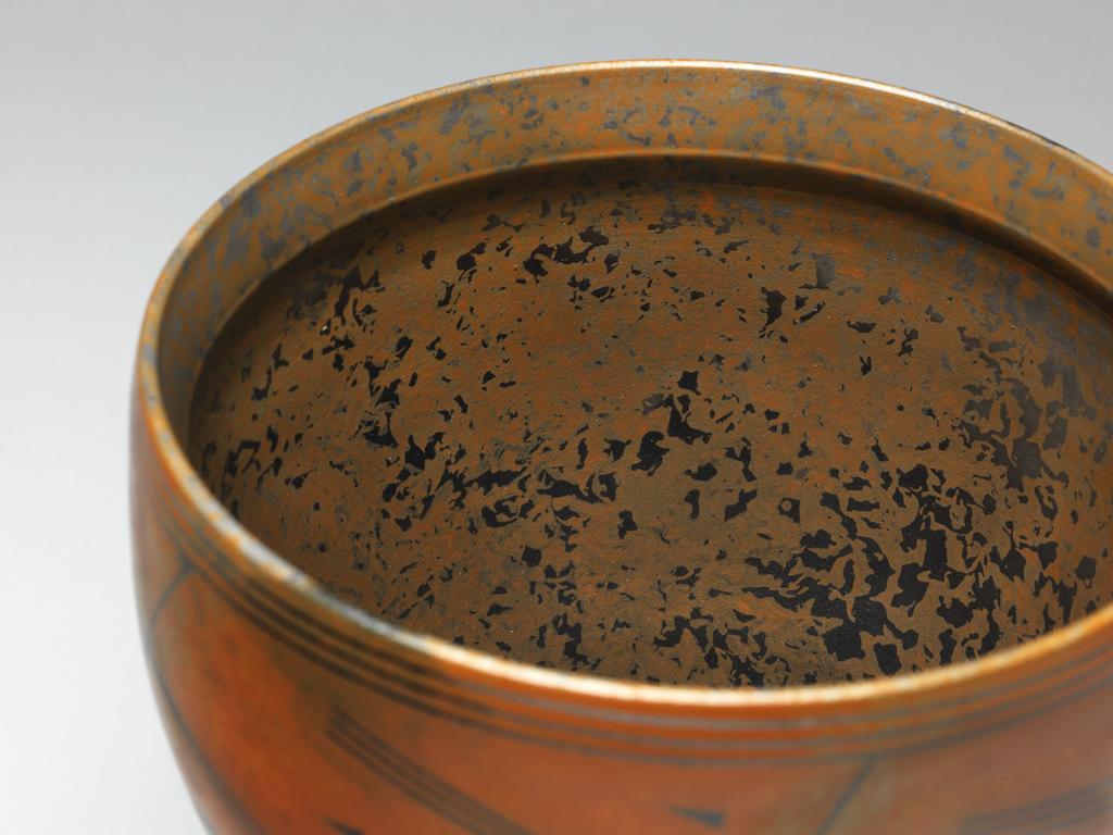 An image of Contemporary Craft. Studio Ceramics. Bowl on Narrow Base. Ross, Duncan (British, b. 1943 India). Circular, standing on a narrow base. The sides slope upwards and outwards to the widest point, and then curve inwards slightly towards the rim. The orange, mottled exterior is decorated round the lower part with pairs of grey broken circles, above which is an abstract design of slanting crosses with pointed triangles in the spaces. There are three dark grey bands round the rim. The interior is mottled in black, grey, and orange. Red earthenware, thrown, coated with terra sigillata slip, decorated with a linear design, burnished, bisque-fired, and smoke fired. Height, whole, 20.5 cm, width, whole, 18.2 cm, 1997. Gift of Nicholas and Judith Goodison through the National Art Collections Fund.
