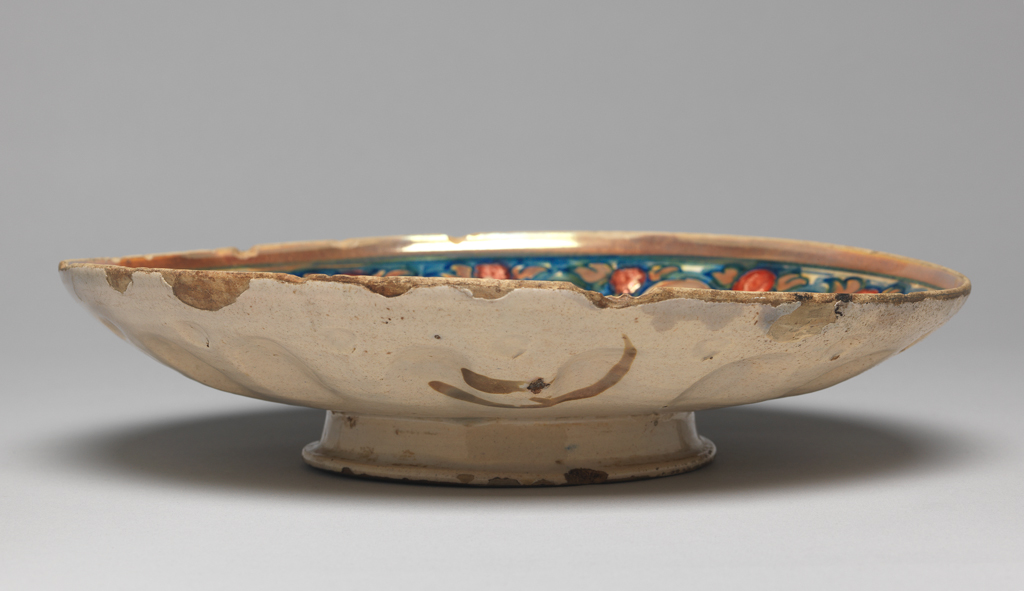 An image of Maiolica shallow bowl standing on a low foot. Coppa. Maestro Giorgio Andreoli, workshop, Umbria, Gubbio. Circular with shallow curved sides and convex boss surrounded by a rib, standing on a low foot. In the middle, within a circular lustred rib, the Agnus Dei, bearing a pennant with a red cross, stands on a red and white chequered pavement. Behind him is a blue skyline and blue striated sky with lustre strokes on it. On the sides, radiating oval leaves alternate with stylised plants bearing spherical buds reserved in a blue ground. A lustre band encircles the rim. The back is decorated in coppery-gold lustre with a stroke in the middle, and three swirls on the sides. Buff earthenware, moulded with decoration in relief and tin-glazed pale beige overall. Painted in blue, and with red and coppery-gold lustre, height, whole, 5.8 cm, diameter, whole, 24.7 cm, circa 1530-1550. Renaissance. Production Notes: The Agnus Dei (Lamb of God) is one of several stock motifs found on the central medallions of Gubbio moulded dishes (coppe), whose sides are decorated with a variety of radiating relief motifs such as leaves, buds and rays.