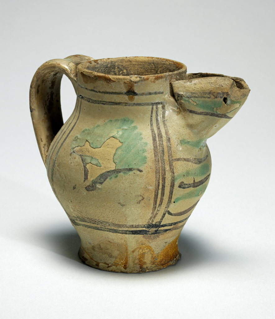 An image of Tin-glazed earthenware jug. Maiolica arcaica. Painted in green and brown with panels, leaves and stripes. Cream earthenware, remnants of lead glaze on lower part and interior, the rest tin-glazed, painted in green and manganese-brown, height 10.3cm, diameter (base) 5.5cm, width (body) 8.8cm, width (handle to spout) 11.4cm, circa 1250-1300. From Umbria, Italy. Medieval.