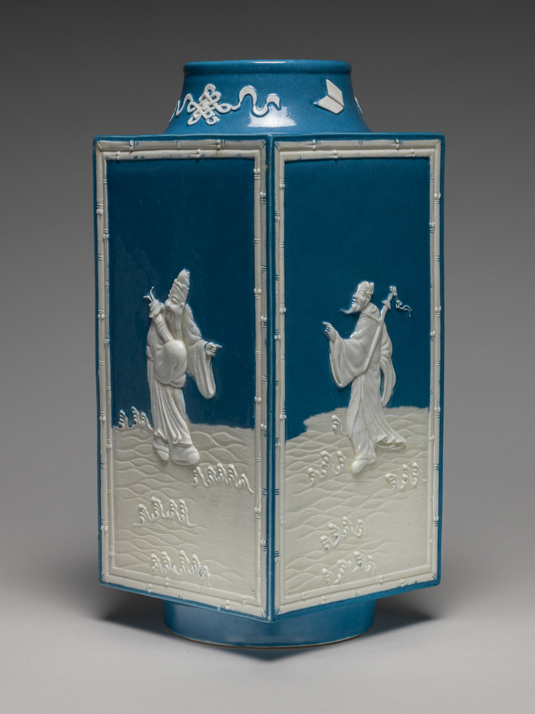 An image of Vase. One of two square stoneware vases, with a sloping cylindrical neck and cylindrical foot ring. With applied white reliefs and opaque powder blue glaze on the interior and parts of the exterior. Decorated on the sides with four of the Eight Immortals standing on waves, enclosed by a rectangular bamboo border, and on the neck with four symbols, all in white. The immortals are Lu Dong-bin (with sword), Li Tie-guai (with crutch and gourd), Lan Cai-he (with basket of flowers) and Zhang Guo-lao (with rods in cane). Mark: Six character seal script mark of Qianlong. Stoneware, powder blue glaze, with applied relief, height 28.5 cm, width 12.5 cm. Qianlong Period (1736-1795). Chinese.