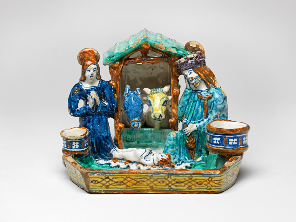 An image of Maiolica. Nativity Inkstand. Giovanni di Nicola Manzoni dal Colle (active early 16th century, Italian, Faenza, circa 1507-1516). With a Nativity group in the round at the back, and an inkwell and candlesocket on either side. The Virgin Mary and St Joseph kneel on either side of a wattle stall occupied by an ox and an ass. The infant Christ lies on a wavy-edged cloth between them. In front of the figures is a space for pens, enclosed by a low wall, decorated on the exterior with strapwork. Dark buff earthenware, tin-glazed on the upper surfaces; base streaked with glaze. Painted in dark blue, green, yellow, brownish-orange, and manganese-purple high-temperature (metallic oxide) colours, height, whole, 15.8 cm, length, whole, 21.5 cm, depth, whole, 17.1 cm, circa 1509-1510. Renaissance.