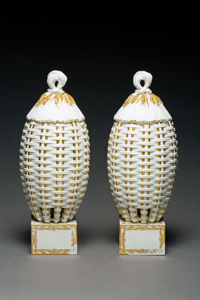 An image of Vase and cover. Beehive Vase (vase ruche no. 1). Sèvres Porcelain Manufactory. The vase is in the shape of a slender beehive made of wickerwork, pierced at the base. The cover is moulded to resemble thatch and has a ring handle in the shape of a doubled over sheaf of corn, tied at the junction with the cover, so that ears of corn radiate from it. Two of each group of three vertical osiers on the vase, the corn stalks and the edges of the thatch on the cover are gilded. The vase stands on a separate square base, decorated with gold scrolls round the outer edges. Glazed soft-paste porcelain beehive vase and cover, decorated with gilding, height, whole, 24.7 cm, height, base, 5.1 cm, 1768-1769. Possibly earlier; the bases were added at an unknown later date. One of a pair.