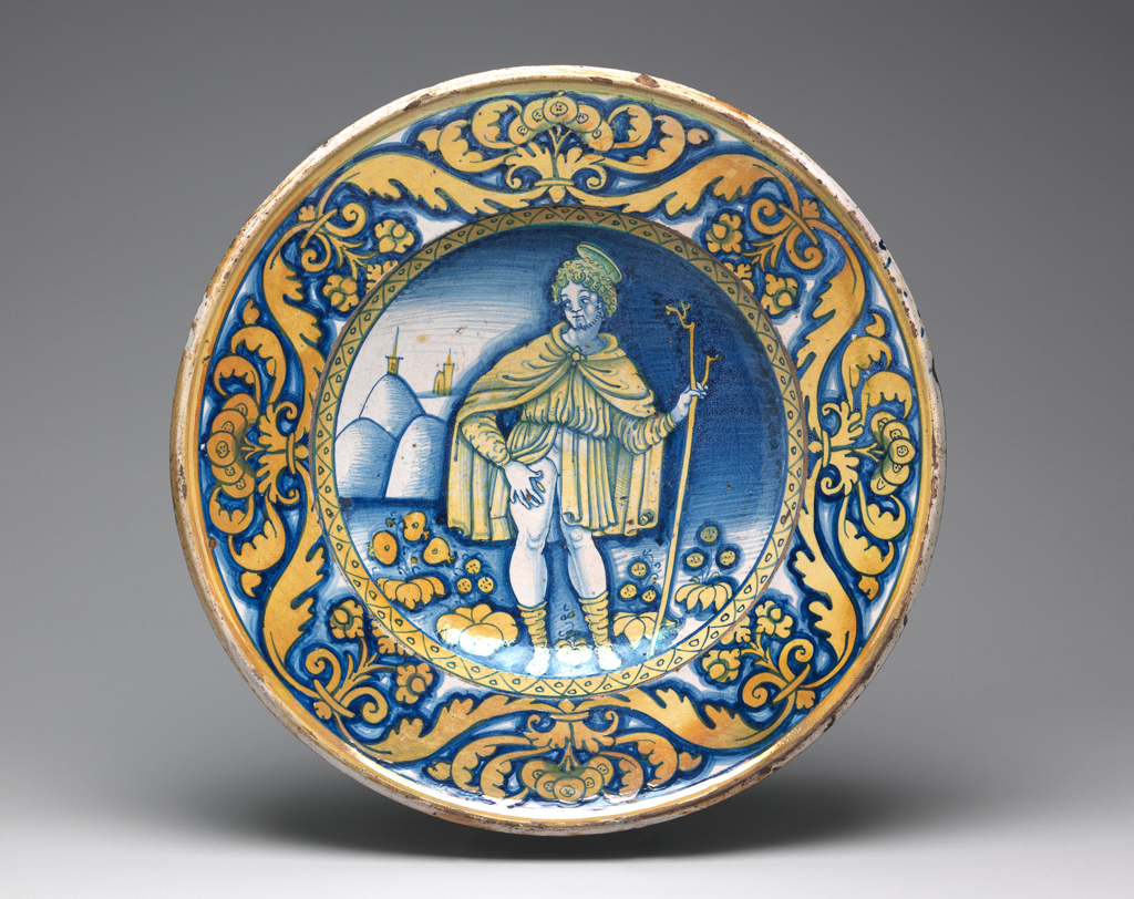 An image of Maiolica. Dish/piatto di pompa. St Roch. Unknown maker, Umbria, Deruta. Shape 61. Circular with slightly sloping rim and wide deep well. standing on a footring pierced by two holes in the correct position for suspension the right way up. In the middle, St Roch leans on a forked staff held in his left hand and points with his right to a plague spot on his right thigh; he has a halo and wears knee-length hose, a short tunic with long sleeves and a cloak. In the background to left, are hills and the towers and spires of two churches, and in the foreground, stylized flowers. The edge of the well has a border of zig-zags and spots. On the rim, there are stylized flowers and scrolling leaves. A band of yellow lustre encircles the outer edge. Dark buff earthenware, the front is tin-glazed; the reverse lead-glazed honey-brown. Painted in bright dark blue and with brassy-yellow lustre, height, whole, 9.0 cm, diameter, whole, 41.8 cm, circa 1500-1550. Renaissance.