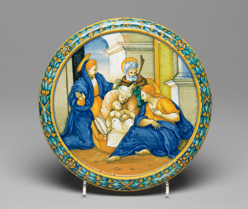 An image of The Holy Family. Maiolica cover from an accouchement set bowl, painted in polychrome with the Holy Family. Tagliere. Milan Marsyas Painter, possibly, The Marches, Urbino. Xanto, Francesco, painter, possibly, The Marches, Urbino. Caraglio, Giovanni Giacomo, printmaker, after. Parmigianino (Francesco Mazzola), painter, after, (Italian, 1503-1540). Circular with a flange on the underside; the upper surface flat with a convex rim. The Holy Family in an architectural setting; on the rim, bound leaves and berries between concentric yellow bands. On the back, two putti standing on clouds support a shield bearing the arms sable, a fess or, a chief party per pale gules and argent two rosettes counterchanged flanked by the letters `ELI' and `PYA'; below, a winged putto's head. Pale buff earthenware, tin-glazed overall. Painted in blue, turquoise-green, yellow, orange, black, grey, and white, height, whole, 1.9 cm, diameter, whole, 19.2 cm, circa 1531. Renaissance. Production Note: If not painted by the 'Milan Marsyas Painter' this was probably painted by Francesco Xanto Avelli da Rovigo. The Holy Family on the top of the cover was derived from the Adoration of the Shepherds, engraved by G.G. Caraglio after Parmigianino.