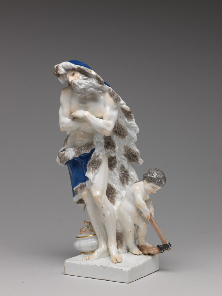 An image of Figure. Winter from a set of Seasons. Meissen Porcelain Manufactory, Saxony, Meissen. Eberlein, Johann Friedrich, modeller (German, 1696-1749). Winter stands with his left knee relaxed and his arms crossed in front of his chest, leaning forward slightly. He has a long grey beard, and wears a blue cloak lined with brown and white fur, which falls down from his head to the ground at the back and is held up across his loins and upper legs in his right hand. On his right, projecting partly over the edge of the base, there is a covered bowl with a fire on top of it, and on his left, is a nude, brown-haired putto cutting wood with an axe. Hard-paste porcelain, press-moulded, glazed, and painted in blue, red, flesh pink, pale and dark brown, grey, and black enamel colours, and lightly gilt on the edges of the bowl only, height, whole, 26.4 cm, width, base, 9 cm, width, whole, 14.6 cm, circa 1745.