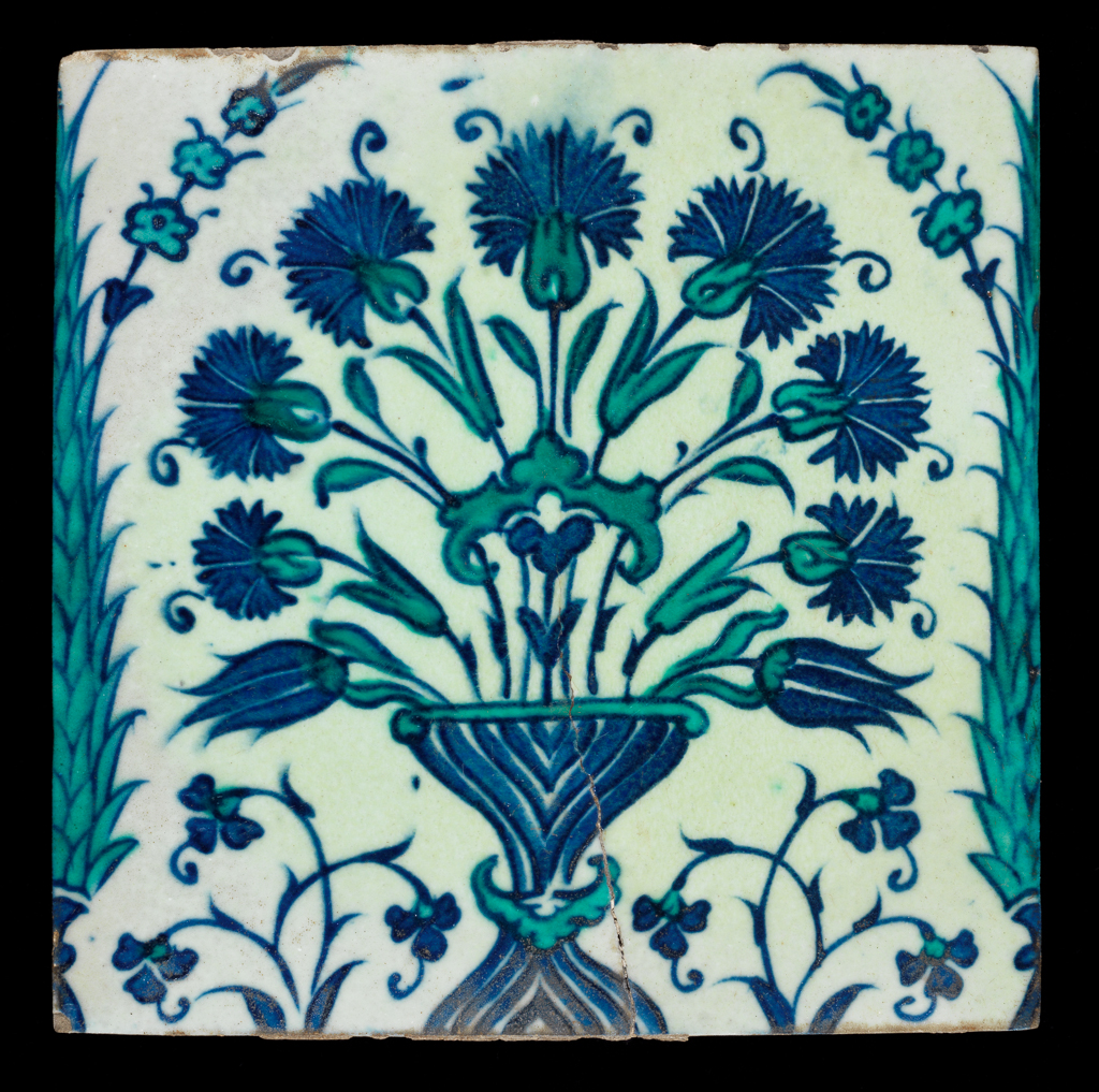 An image of Fritware/Stonepaste Tile. Unknown. Upper side: a spray of blue carnations spring from a blue vase, both the spray and vase are clasped by a green agrafe. Either side of the vase, two smaller floral sprays and along each vertical edge two partial cypress trees with sprouting prunus style branches, all painted in blue and green. Probably fritware, mould made, painted under the glaze in blue and green under a colourless glaze, height 25.0 cm, width 25.2 cm, depth 1.0 cm, weight, whole, 1191 g, circa 1600-1799. Ottoman Period. Turkey, probably Iznik.