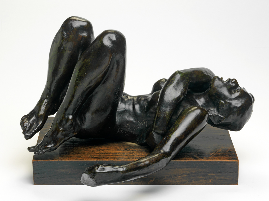 An image of Sculpture/Figure. Une Damnée. Rodin, Auguste (French, 1840-1917), Paris, Colmar. Bronze, cast, with dark brown patina. The woman lies on her back with her legs drawn up, her left arm extended to the side, and her right arm across her body with the hand touching her left side. Her head is tilted back and her mouth is wide open. The figure is attached by means of a screw to a rectangular wooden base with a black and brown striped painted surface. Notes: Conceived c. 1884-85 as a study for a figure on the viewer's right side of the Tympanum of The Gates of Hell. Date of cast uncertain, probably 1964-69, when twelve casts were made by Georges Rudier.